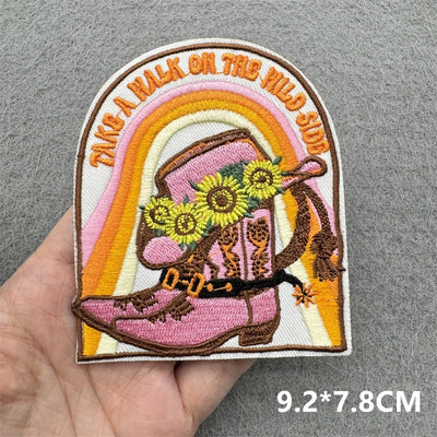 Embroidered patch with pink cowboy boot, flowers, and rainbow text held by a hand on a gray background.