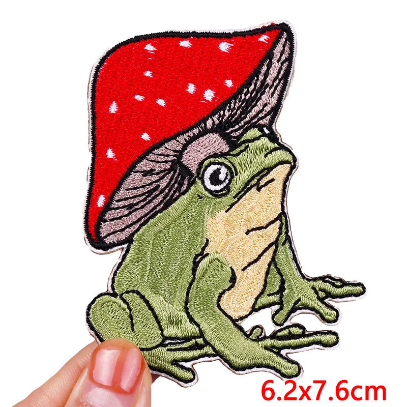 Embroidered patch of a frog with a red mushroom hat held by a hand on a white background.