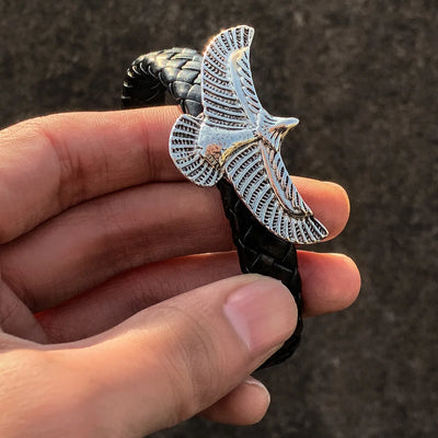 Hand holding a black bracelet with a silver eagle emblem against a dark background