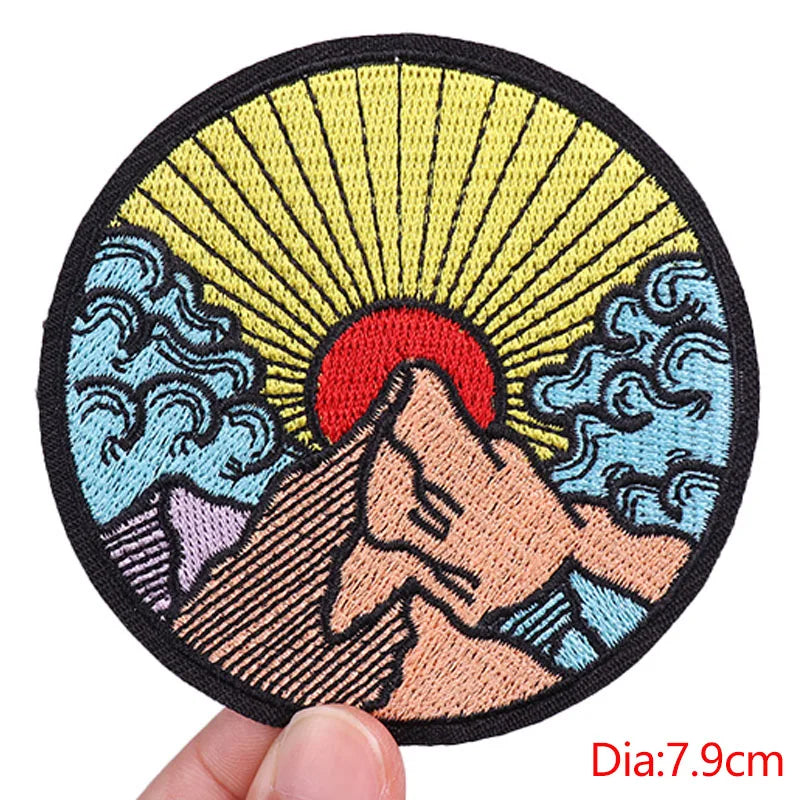 Round embroidered patch with mountain, sun, and waves design held by a hand.