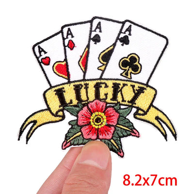 Embroidered patch with playing card symbols and 'LUCKY' banner held by a finger, on a white background.