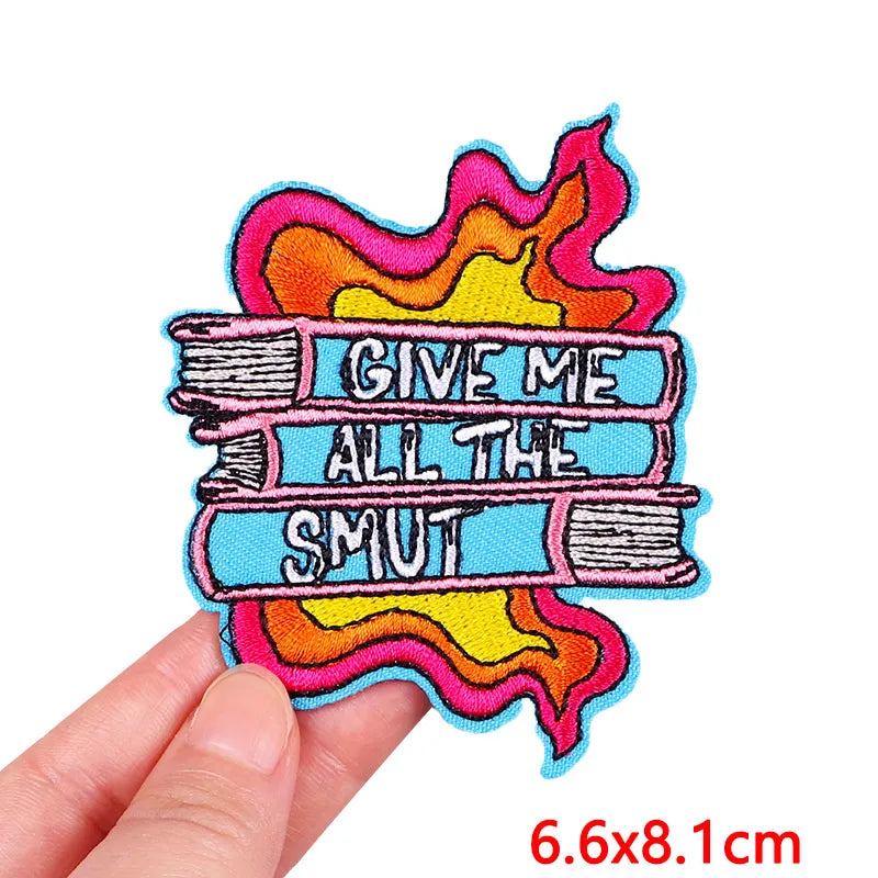 Colorful patch with text 'Give Me All The Smut' held by a hand, on a white background.