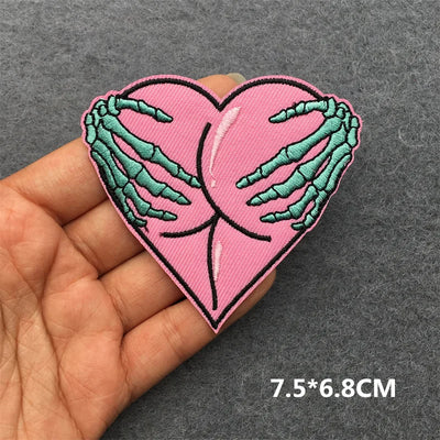 Heart-shaped patch with skeleton hands holding a pink heart, held in a hand against a gray background.
