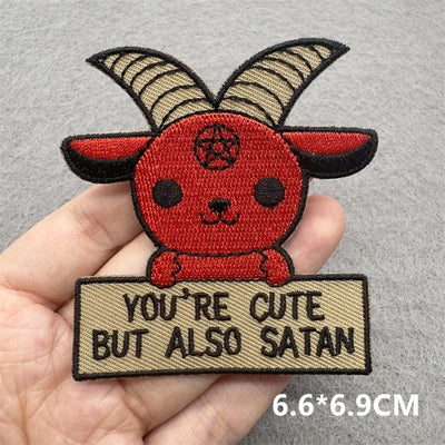 Embroidered patch with a red creature design and text 'YOU'RE CUTE BUT ALSO SATAN' held by a hand on a gray background.