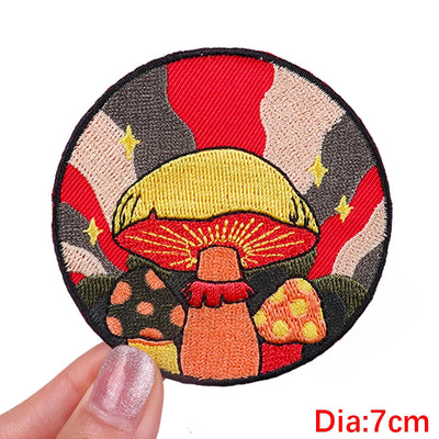 Circular embroidered patch with mushrooms held by a hand, on a white background.