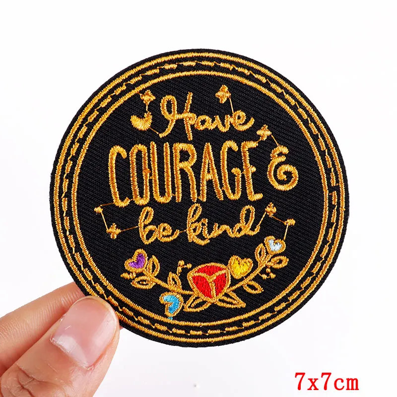 Hand holding a black gold red purple and blue embroidered Have Courage and Be Kind iron-on patch approximately 7x7cm on a white background