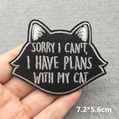 Black cat-shaped patch with text 'Sorry I can't, I have plans with my cat' held in a hand against a gray background.