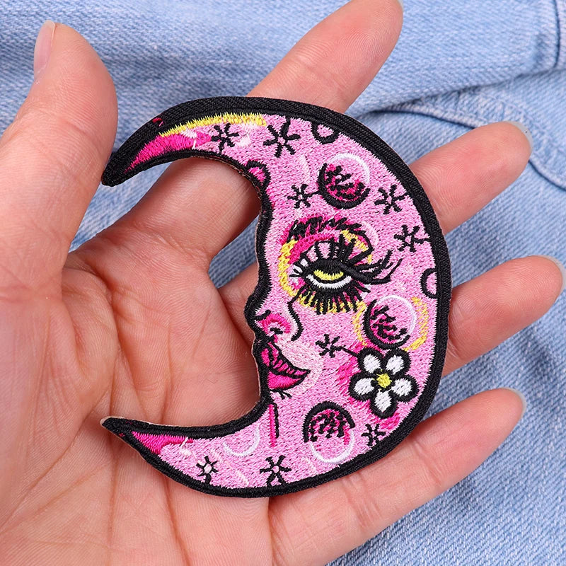 Hand holding a pink crescent moon patch with face design against a denim background