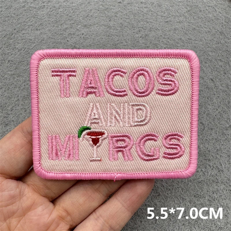 Hand holding a pink and cream embroidered Tacos and Margs iron-on patch with margarita glass accent approximately 5.5x7cm on a gray background
