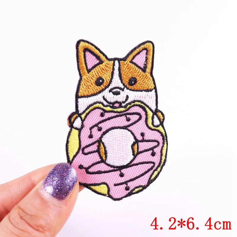 Embroidered patch of a dog holding a donut, held by a hand with glittery nail polish on a white background.
