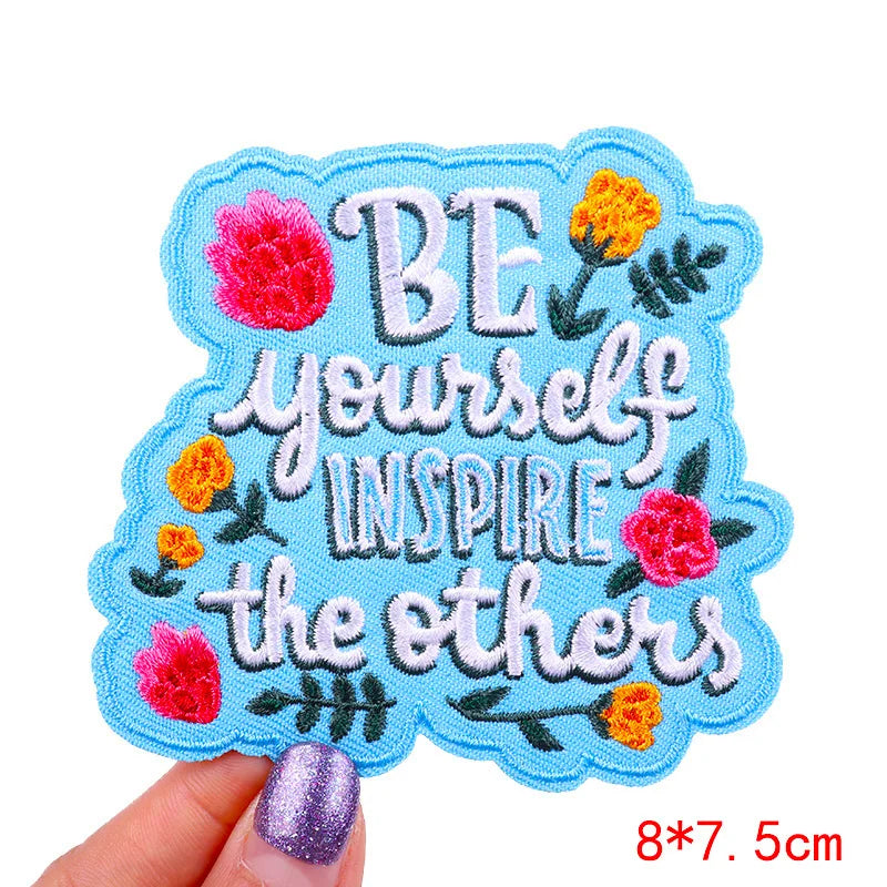 Embroidered patch with 'Be yourself, inspire the others' text and floral designs held by a hand.