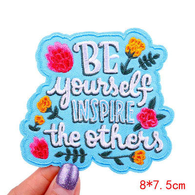 Embroidered patch with 'Be yourself, inspire the others' text and floral designs held by a hand.
