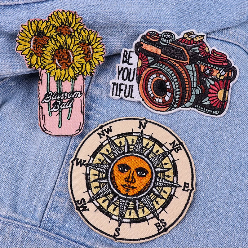 Three embroidered patches on a denim jacket: sunflowers in a jar, camera with 'Be You Tiful' text, and compass design.