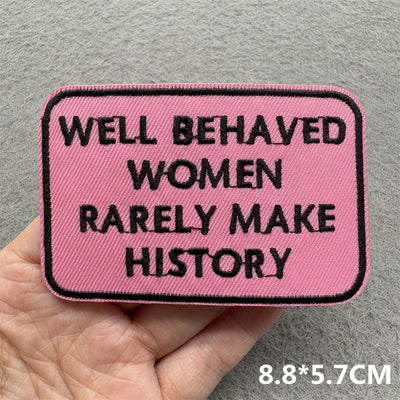 Pink patch with text 'Well Behaved Women Rarely Make History' held in a hand against a gray background.
