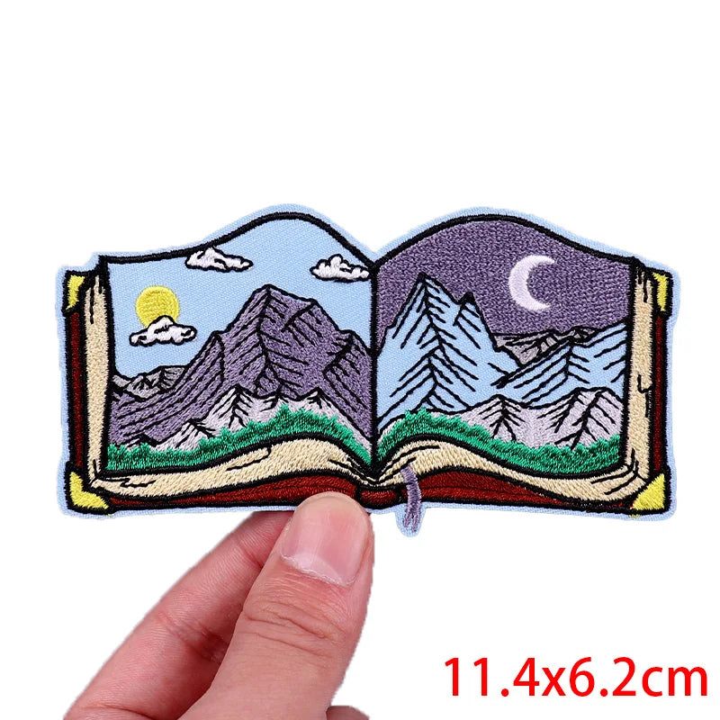 Embroidered patch of an open book with mountain scenes held by a hand, with dimensions displayed.