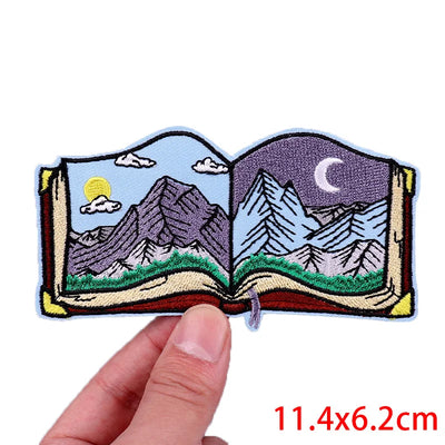 Embroidered patch of an open book with mountain scenes held by a hand, with dimensions displayed.