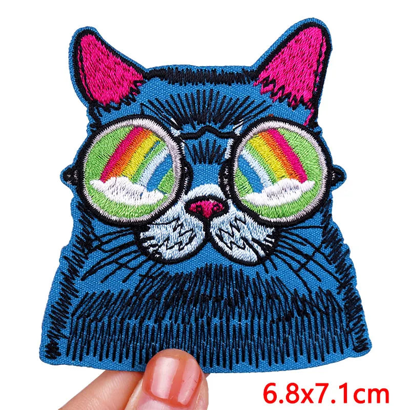 Embroidered patch of a blue cat with rainbow glasses held by a hand, on a white background.