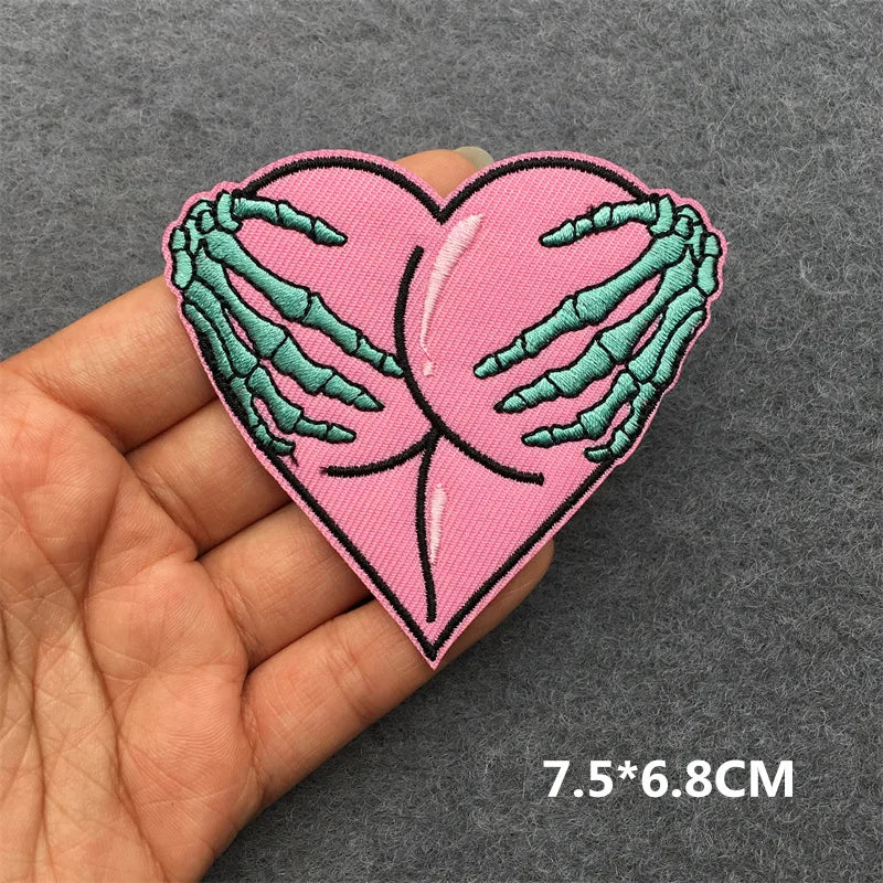 Heart-shaped patch with skeleton hands holding a pink heart, held in a hand against a gray background.
