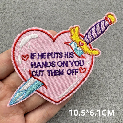 Heart-shaped patch with embroidered text and designs held by a hand on a gray background