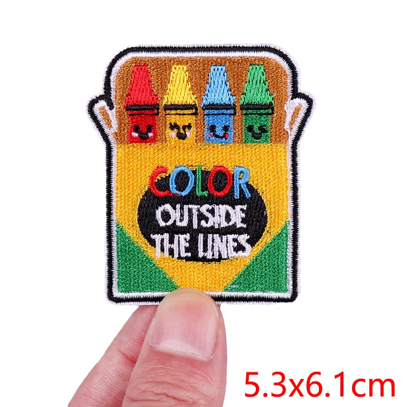 Embroidered patch with crayons and 'Color Outside The Lines' text held by a hand.