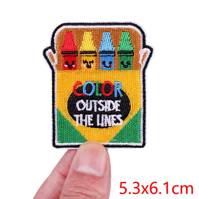 Embroidered patch with crayons and 'Color Outside The Lines' text held by a hand.