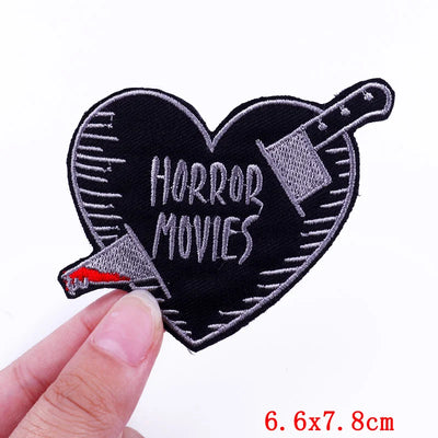 Heart-shaped patch with 'Horror Movies' text and a knife design, held by a hand on a white background.