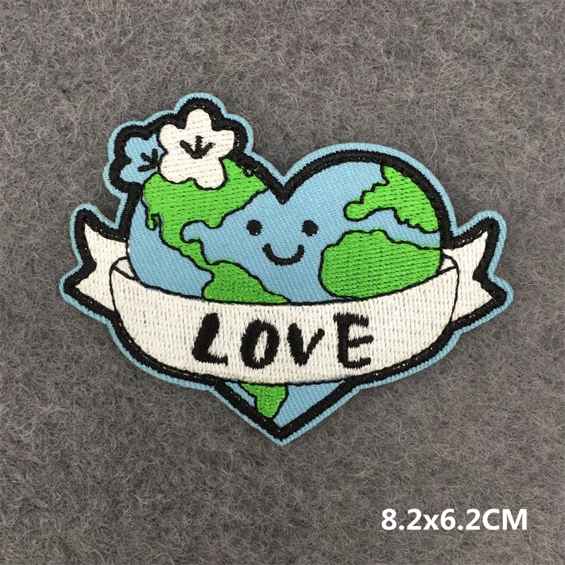 Hand holding a blue green white and black embroidered Earth Love iron-on patch with smiling earth heart and flower design approximately 8.2x6.2cm on a gray background