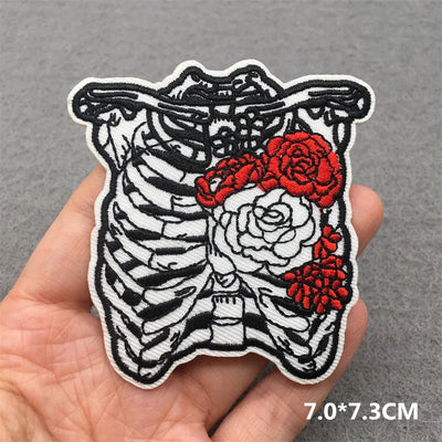Embroidered patch of a skeleton chest with red and white roses held in a hand against a gray background.