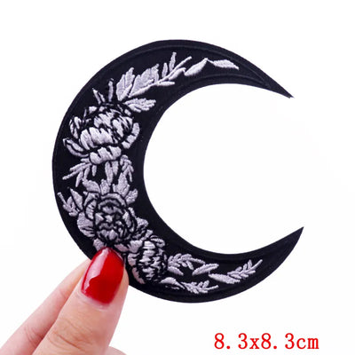 Decorative crescent moon patch with floral designs held by a hand with red nail polish, on a white background.