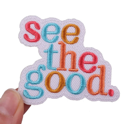 Embroidered patch with 'see the good' text in colorful letters held by a hand on a white background