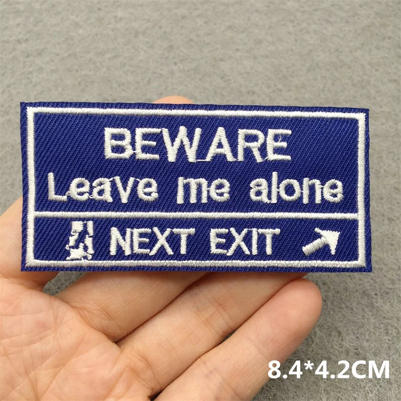 Embroidered patch with text 'BEWARE Leave me alone NEXT EXIT' held by a hand on a gray background.