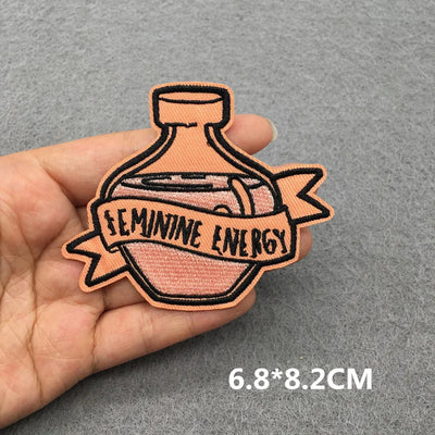 Patch with 'Feminine Energy' text held in a hand on a gray background
