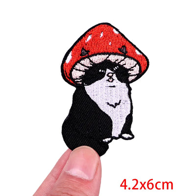 Embroidered patch of a cat wearing a red mushroom hat held between fingers on a white background.