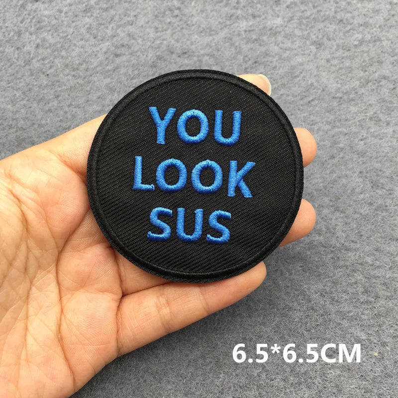 Hand holding a black and blue embroidered You Look Sus iron-on patch approximately 6.5x6.5cm on a gray background