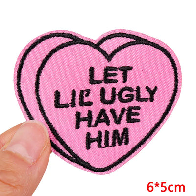 Pink heart-shaped patch with text 'LET LIE UGLY HAVE HIM' held by a hand on a white background.