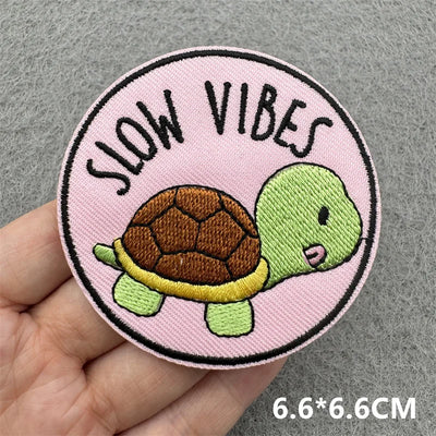 Circular patch with a turtle and 'Slow Vibes' text on a pink background, held by a hand.