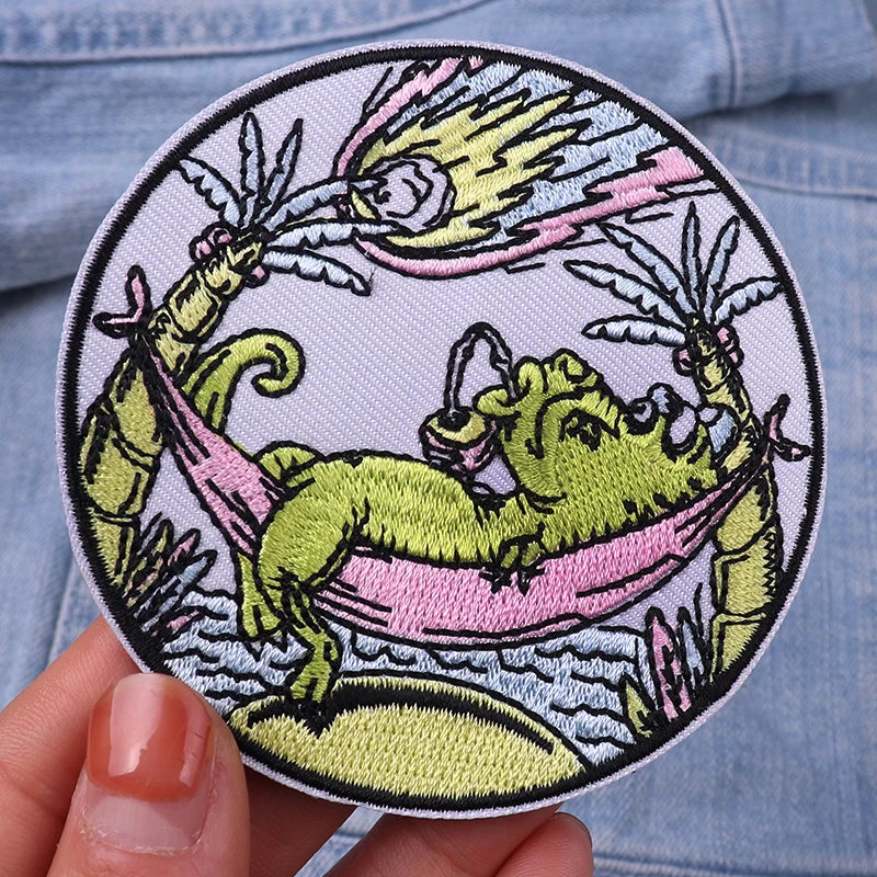 Embroidered patch of a green dragon lounging in a hammock with two crabs, held against a denim background.