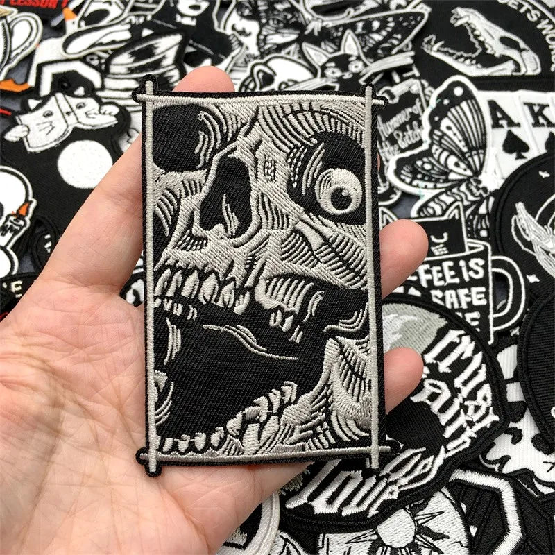 Hand holding a skull-themed patch with a black and white design against a background of various other patches.