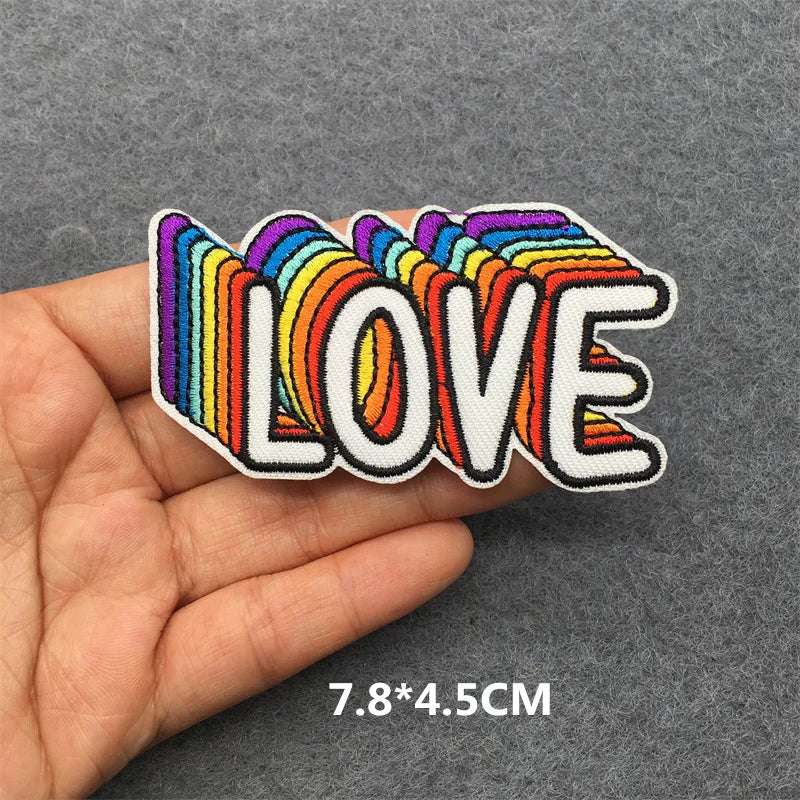Hand holding a rainbow multicolor embroidered LOVE iron-on patch approximately 7.8x4.5cm on a gray background