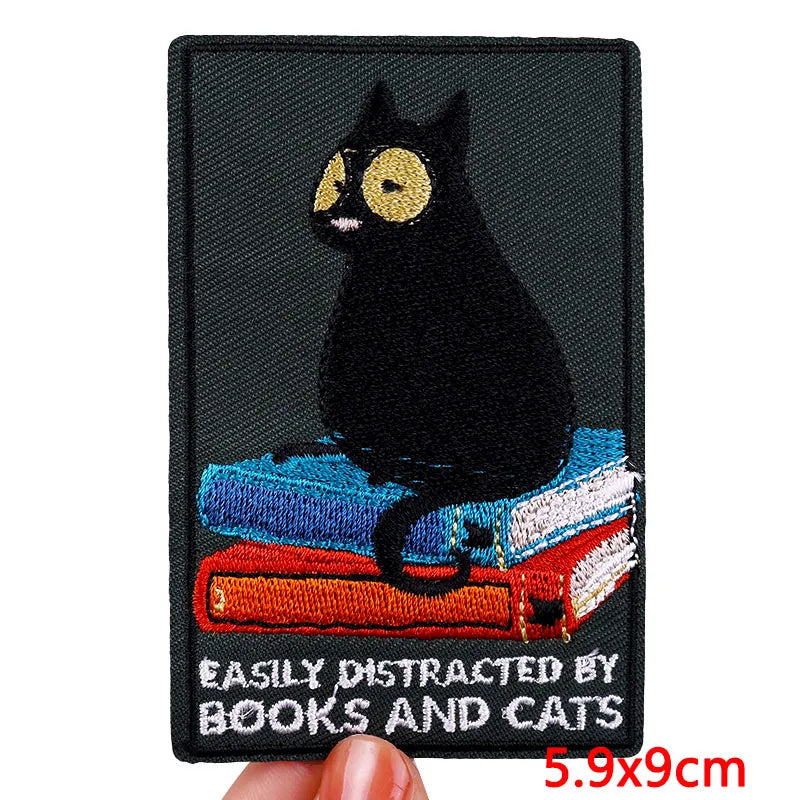 Embroidered patch with a black cat on books and text 'Easily Distracted by Books and Cats' held by a hand.