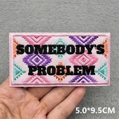 Embroidered patch with 'SOMEBODY'S PROBLEM' text held in a hand against a gray background