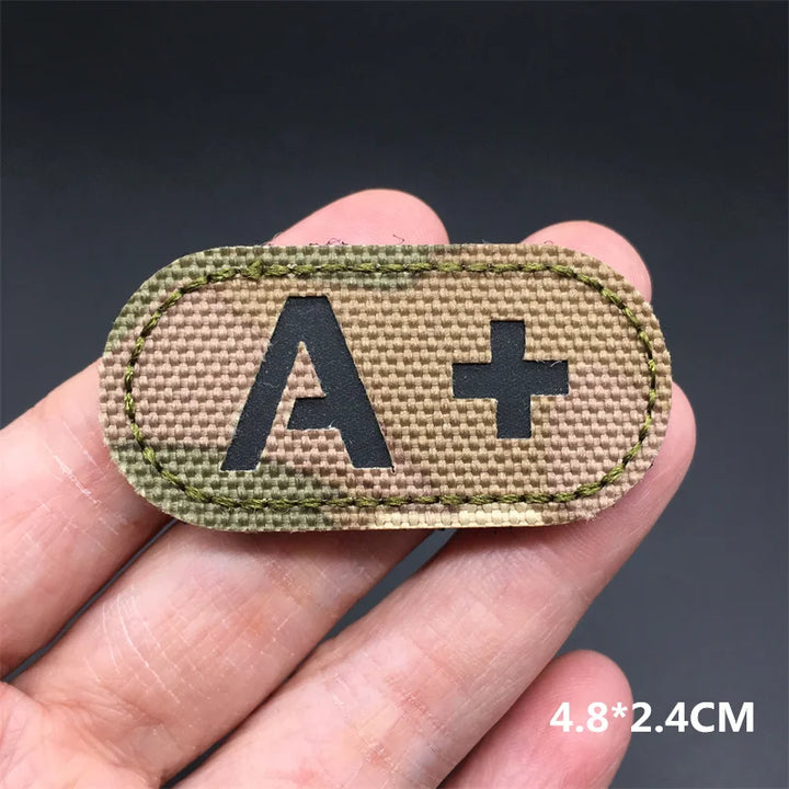 Hand holding an A+ multicam IR reflective tactical blood type hook and loop patch approximately 4.8x2.4cm on a dark background