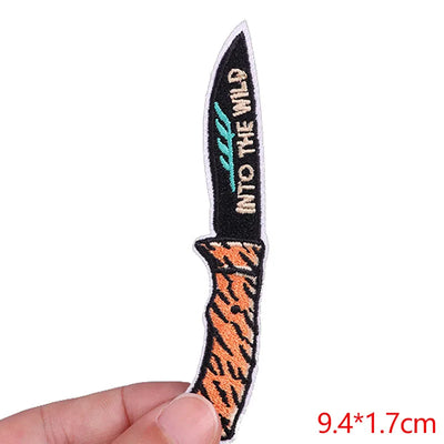 Small embroidered knife patch with 'Into the Wild' text held by a hand on a white background.