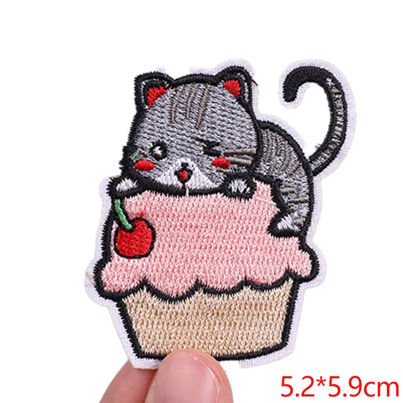 Embroidered patch of a cat on a pink cupcake with a cherry, held between fingers.
