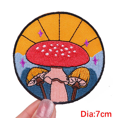 Circular embroidered patch with a red mushroom design held by a hand, against a white background.