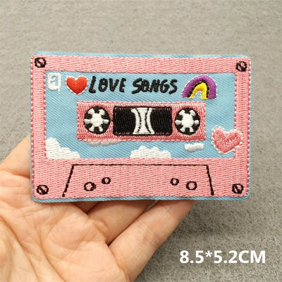 Embroidered patch of a pink cassette tape with 'Love Songs' text held in a hand on a gray background.