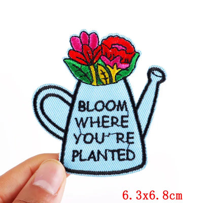 Embroidered patch of a watering can with flowers and 'Bloom Where You're Planted' text, held by a hand.