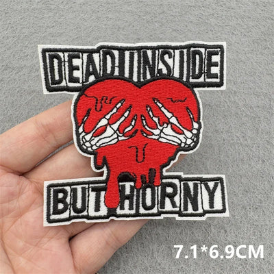 Embroidered patch with skeleton hands holding a red heart and text 'DEAD INSIDE BUT HURRY' held by a hand on a gray background.