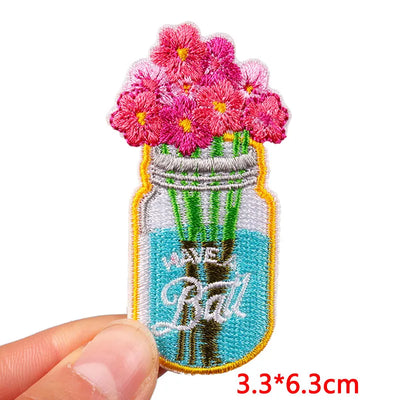 Embroidered patch of a mason jar with flowers held by a hand, on a white background.