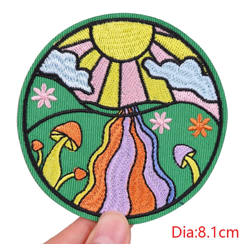 Circular embroidered patch with a sun, mushrooms, and flowers held by a hand on a white background.